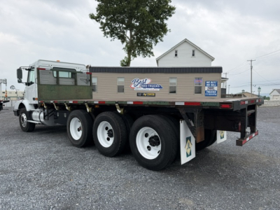 Flatbed truck with green cargo bed and white cab, 3 axles, visible wheels, parked on gravel lot; Volvo 2006 flatbed truck listing image.
