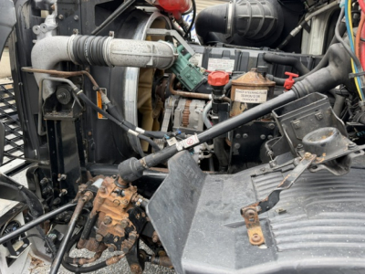Engine bay of a 2006 Volvo VHD flatbed truck showing battery, hoses, alternator, and hydraulic components.