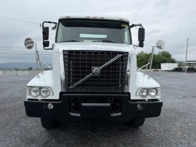 Volvo VHD front view showing large grille with Volvo emblem, dual rectangular headlights, black bumper, and side mirrors; flatbed truck aesthetic.