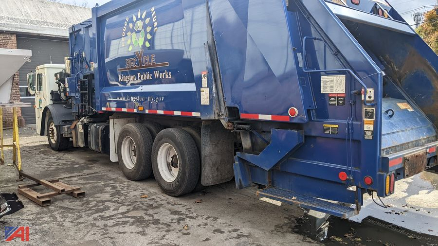Rear view of a blue 2013 Mack LEU 613 trash truck showing refuse body and rear platform with hydraulic components.