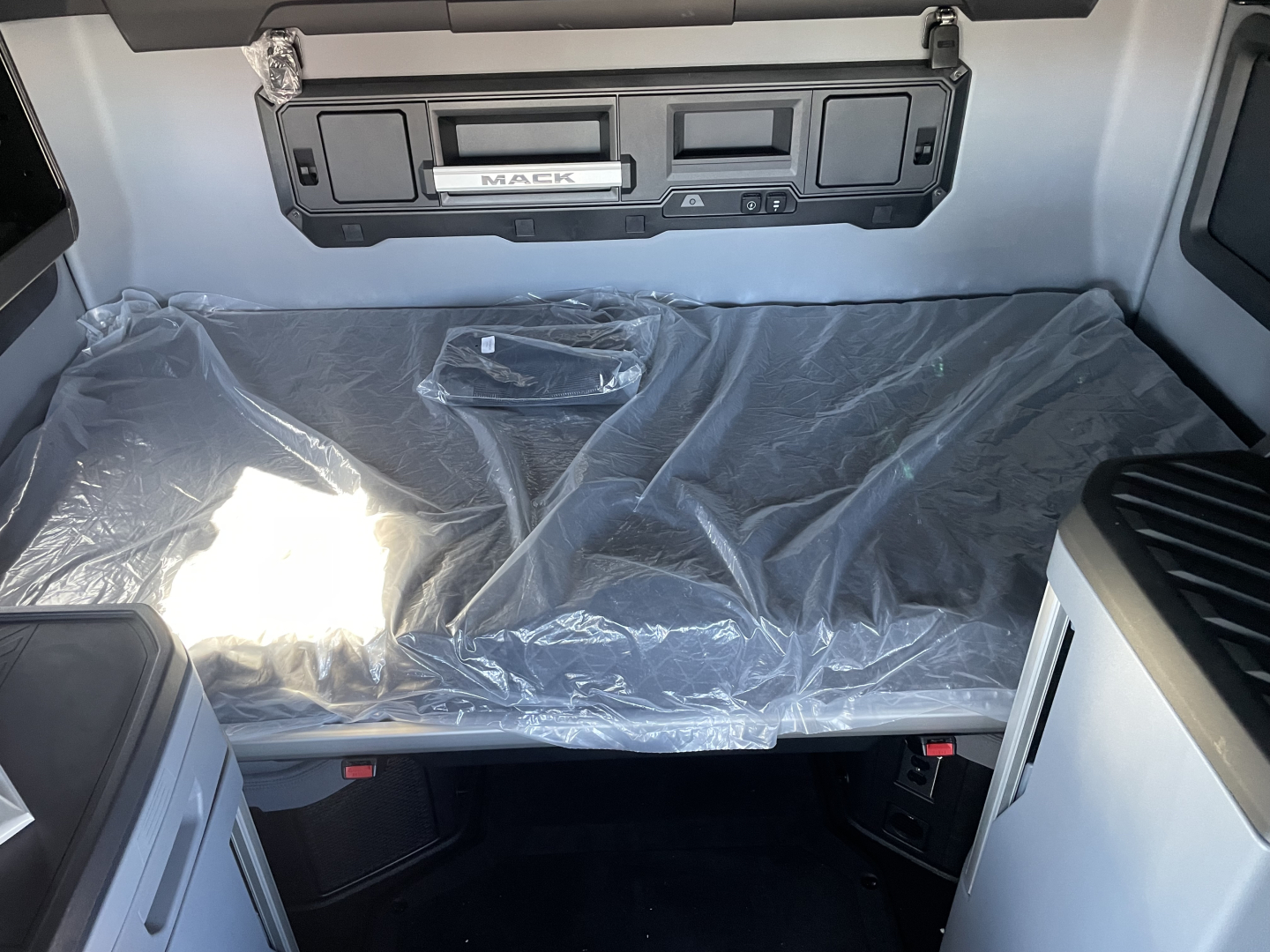 Truck cab interior showing a folded bunk bed covered in plastic, Mack emblem on the overhead console, and control panels in the dash area.