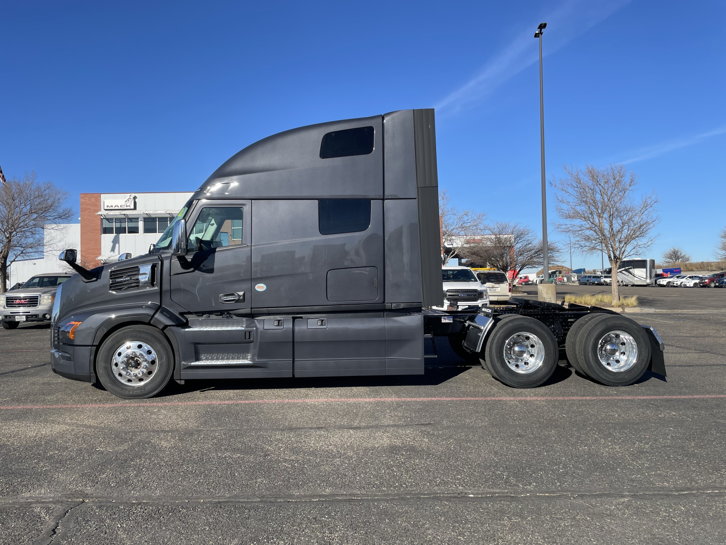 Truck side view showing a black Mack PR64T76H sleeper tractor with three rear axles and chrome wheels.