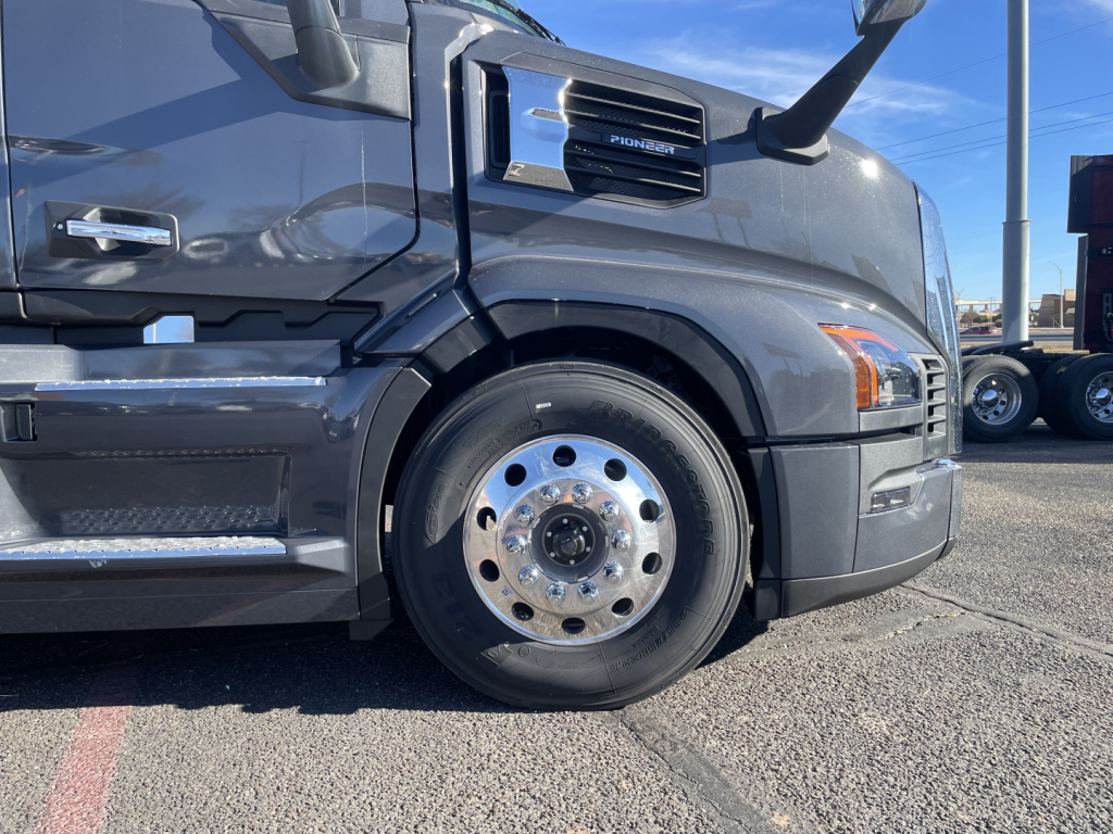 Front left wheel well of a Mack PR64T76H truck showing front fender, headlight area, and polished steel wheel with hub and lug nuts.