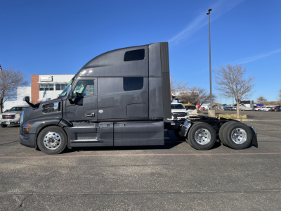 Truck side view showing a black Mack PR64T76H sleeper tractor with three rear axles and chrome wheels.