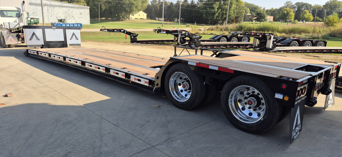 Trailer showing a long flatbed with tandem axles and slide-out mechanism, reflective safety stripes, and dual rear wheels.
