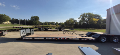 Trailer deck showin g a low-angle view of a double drop detachable trailer with a slide-out section extended, parked on a concrete surface with a white cab and trailer in background.