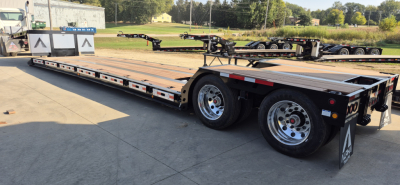 Trailer showing a long flatbed with tandem axles and slide-out mechanism, reflective safety stripes, and dual rear wheels.