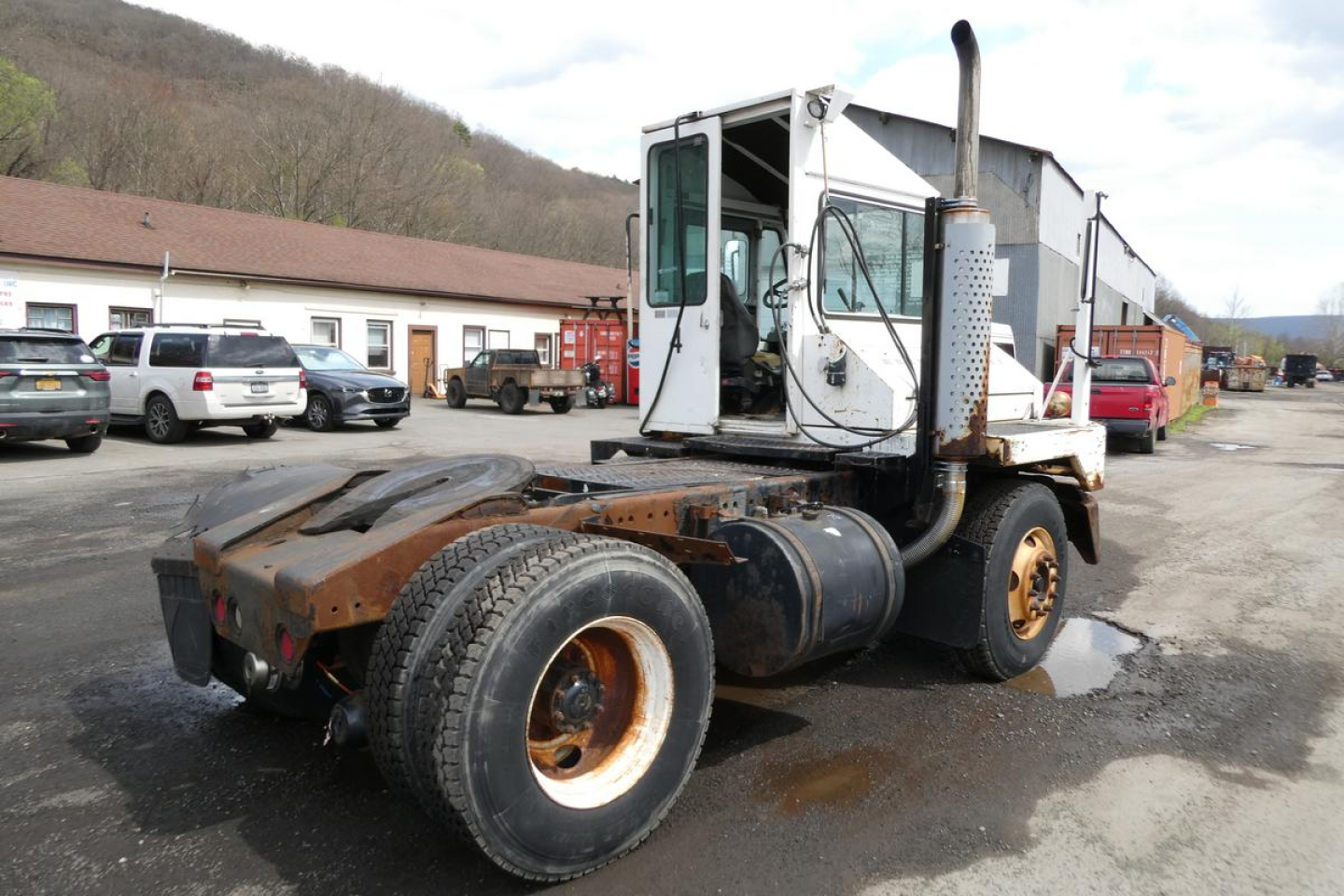 Truck tractor chassis with white cab, exposed exhaust stack, and dual rear wheels; rusty frame and tires visible; background shows a yard with vehicles.