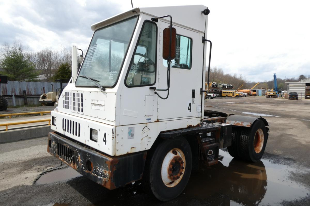 Industrial yard truck cab with white exterior, rectangular windshield, side mirrors, and exposed chassis; visible rust on lower bumper and wheel area.