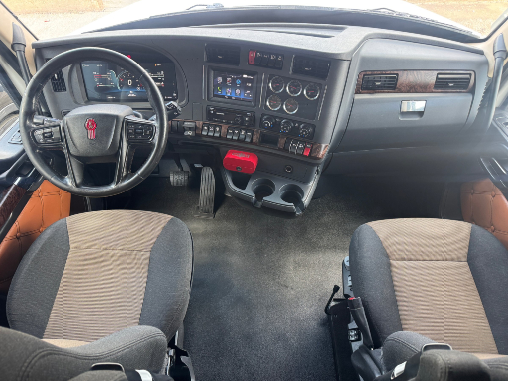 Kenworth truck interior dashboard and seats with steering wheel, center console, and instrument cluster. Visible text includes dashboard controls and a red tag hanging near the center.