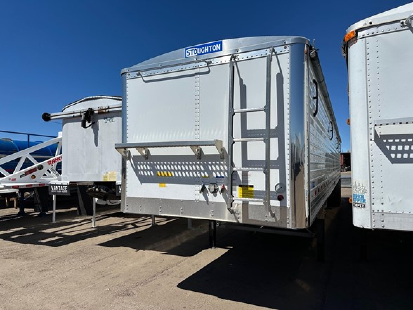 Stoughton air ride grain trailer in a row of trailers, silver aluminum, with rear ladder and loading chute visible.