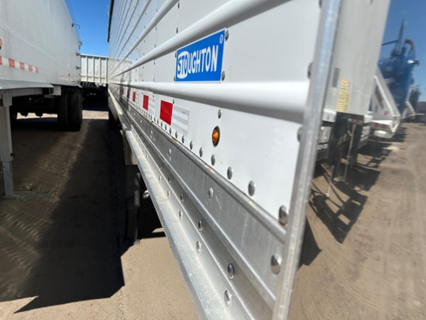 Stoughton air ride grain trailer side showing Stoughton logo and riveted aluminum siding with reflective red/white tape, dirt ground, other trailers in background.