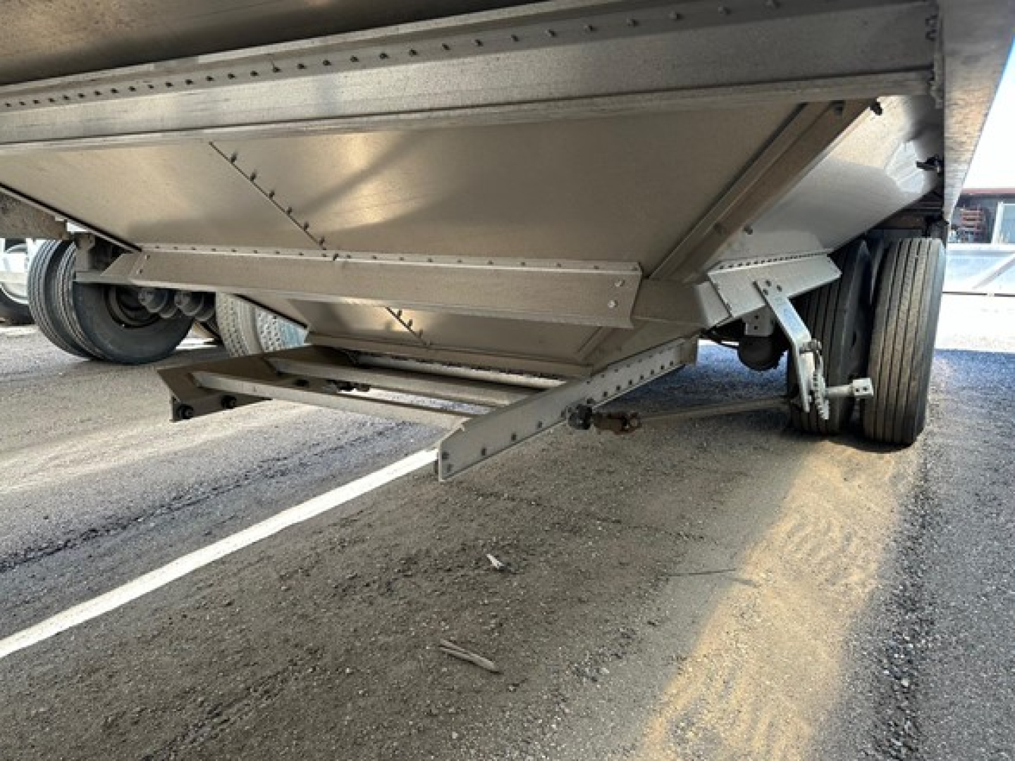 Trailer underside showing hopper grain trailer landing gear and suspension; steel beams, hinges, and a reinforced hopper bottom visible.