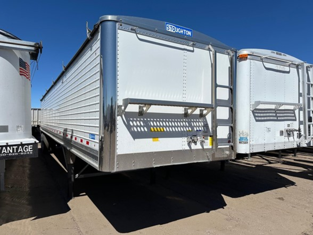 Stoughton air ride grain trailer in a storage lot, silver with blue accents and a Stoughton logo on the front header; hopper trailer with a hinged front door and ladder racks.