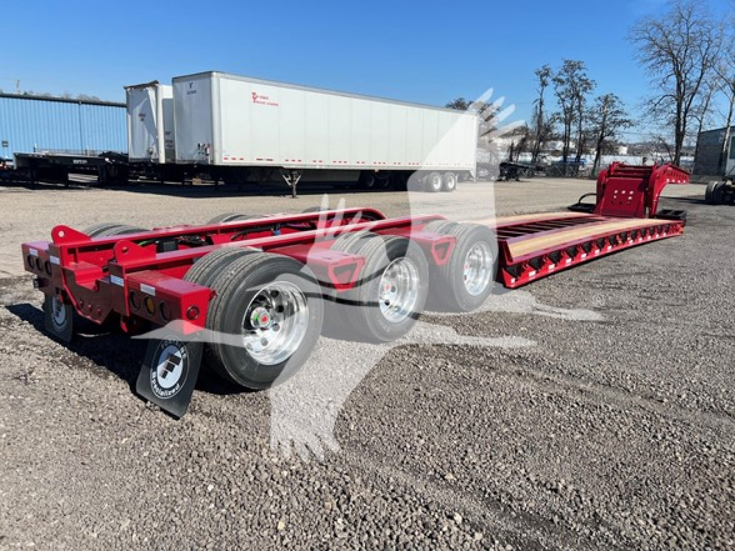 Trailer chassis showing a red Fontaine lowboy step deck with tandem axles and long flatbed, no text visible on side, gravel lot with other trailers in background.