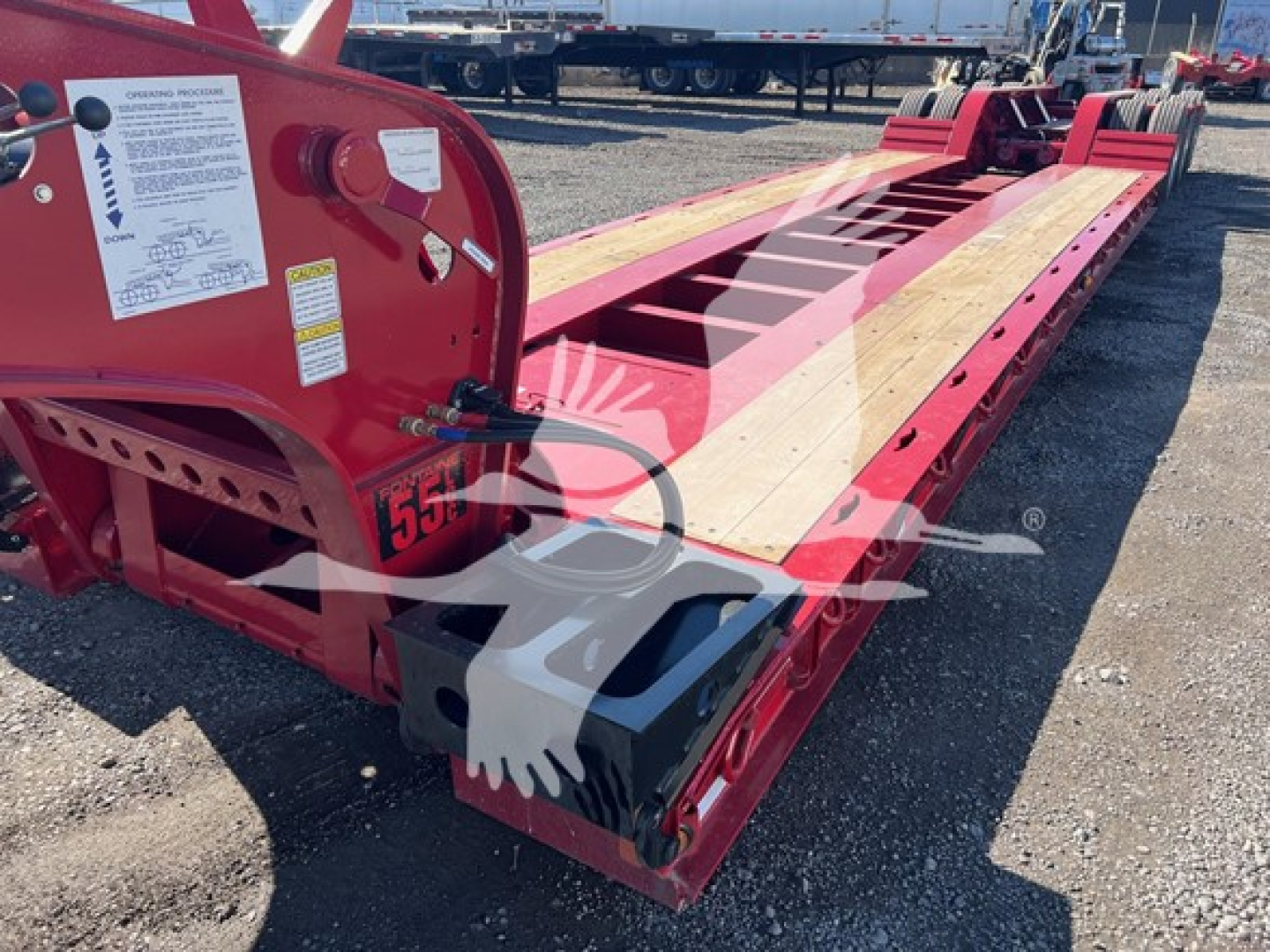 Fontaine lowboy trailer side view showing red frame and wooden deck with black-drive components; label stickers on front left; trailer frame and wheel areas visible.