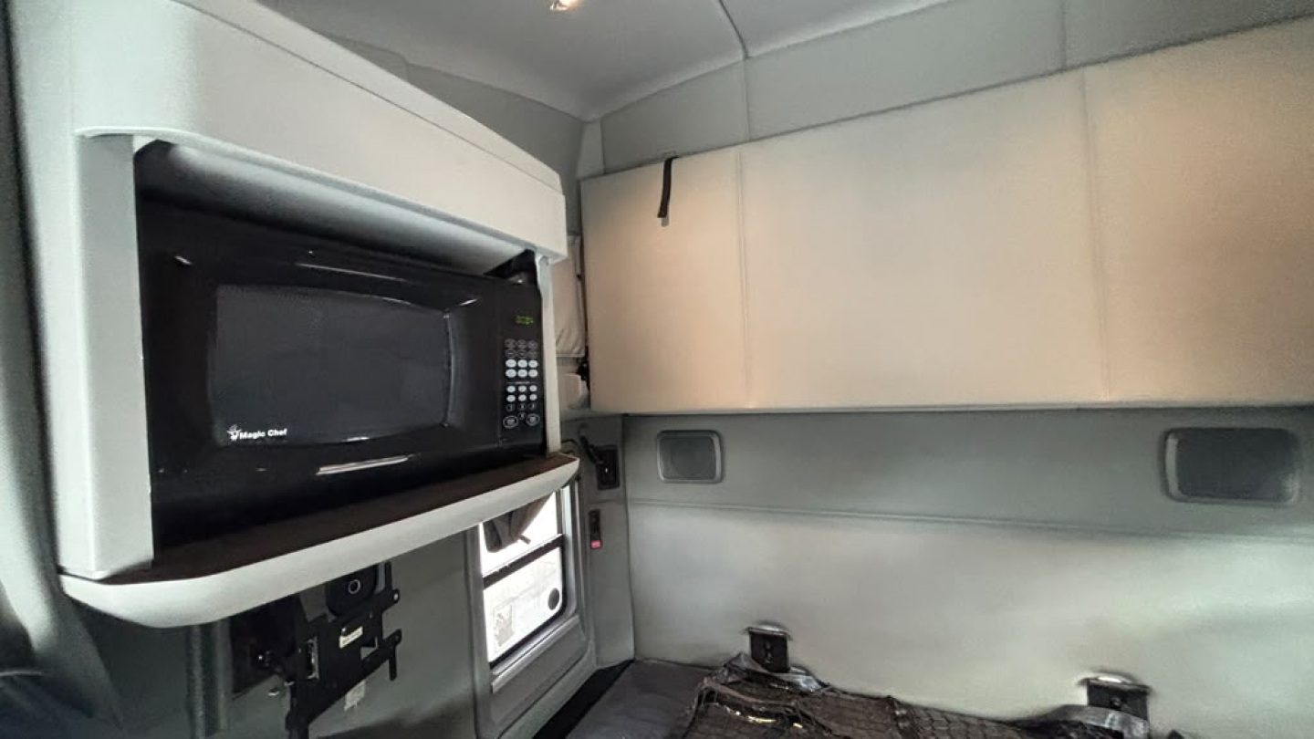 Kenworth T680 sleeper cab interior showing microwave oven mounted above storage with windowed sleeper area and side panel shelves.