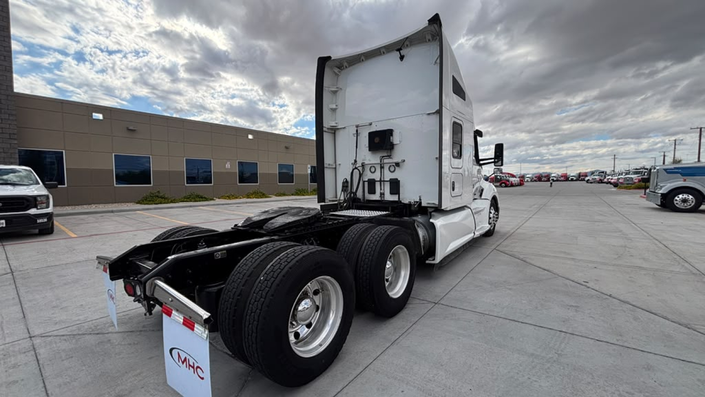 Kenworth T680 tractor with white sleeper, rear chute and dual rear axles, viewed from left-rear, showing fifth wheel area and back of cab. Parking lot with other trucks.