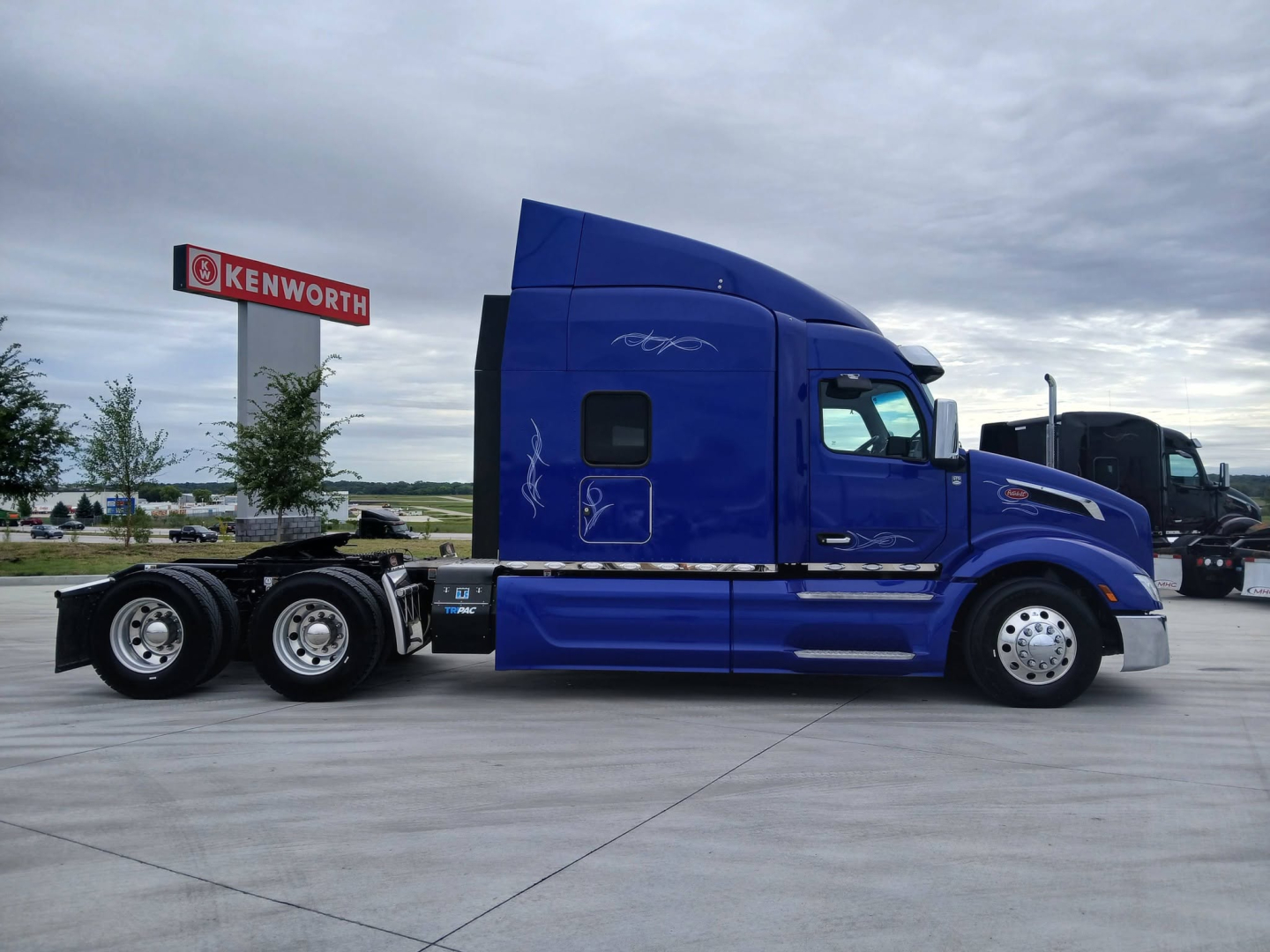 Peterbilt 579 sleepered tractor in blue side profile with triple rear axles, chrome fuel tanks, and Kenworth sign in background.