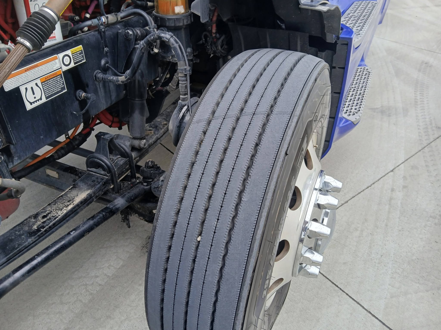 Peterbilt 579 truck front wheel and chassis close-up showing front steering linkage and tire tread, with VIN sticker area and cab area visible.