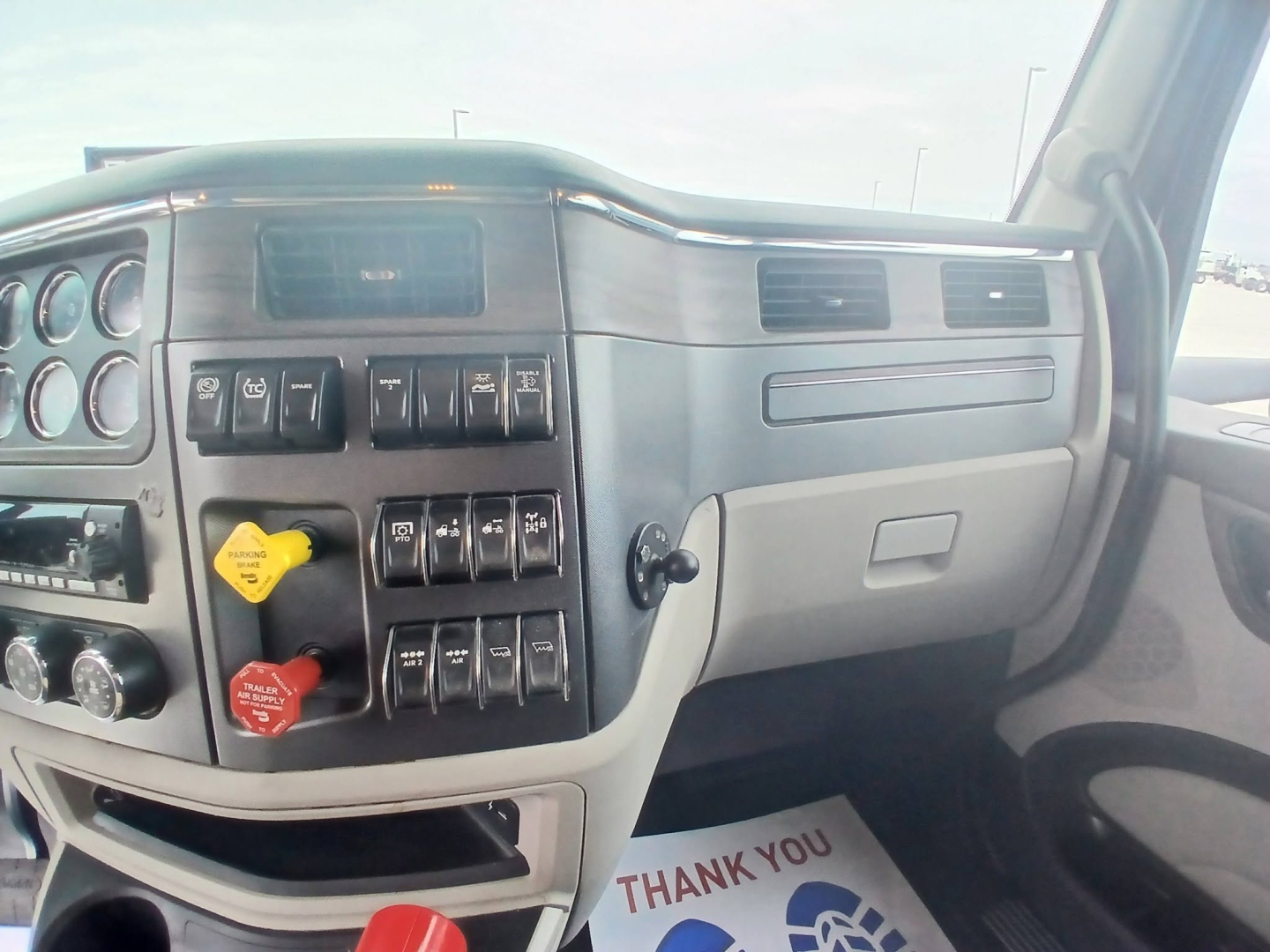 Peterbilt truck interior dashboard with numerous switches and gauges visible; yellow and red control knobs on the left side of the panel; partial glove compartment on the passenger side; windshield and side window; driver’s door frame.