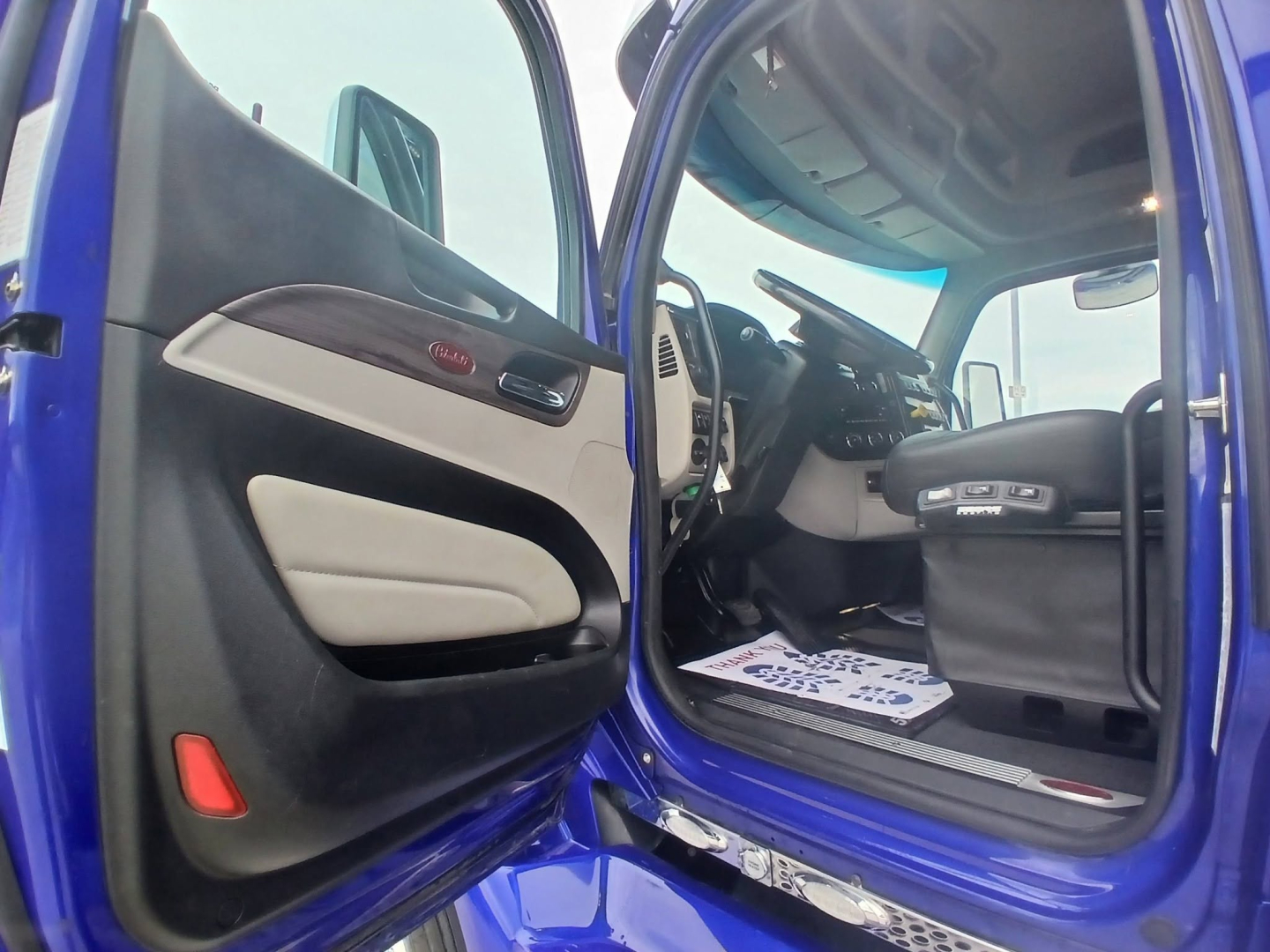 Peterbilt 579 truck interior view through open driver door, blue exterior, beige/black door panels, steering wheel, dashboard, seats visible.