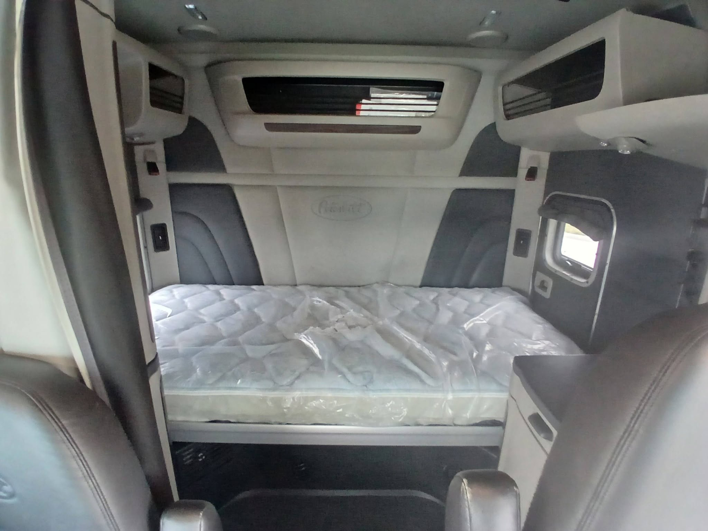 Interior of a Peterbilt 579 sleeper cab showing bed with mattress, overhead storage, and cabin windows.