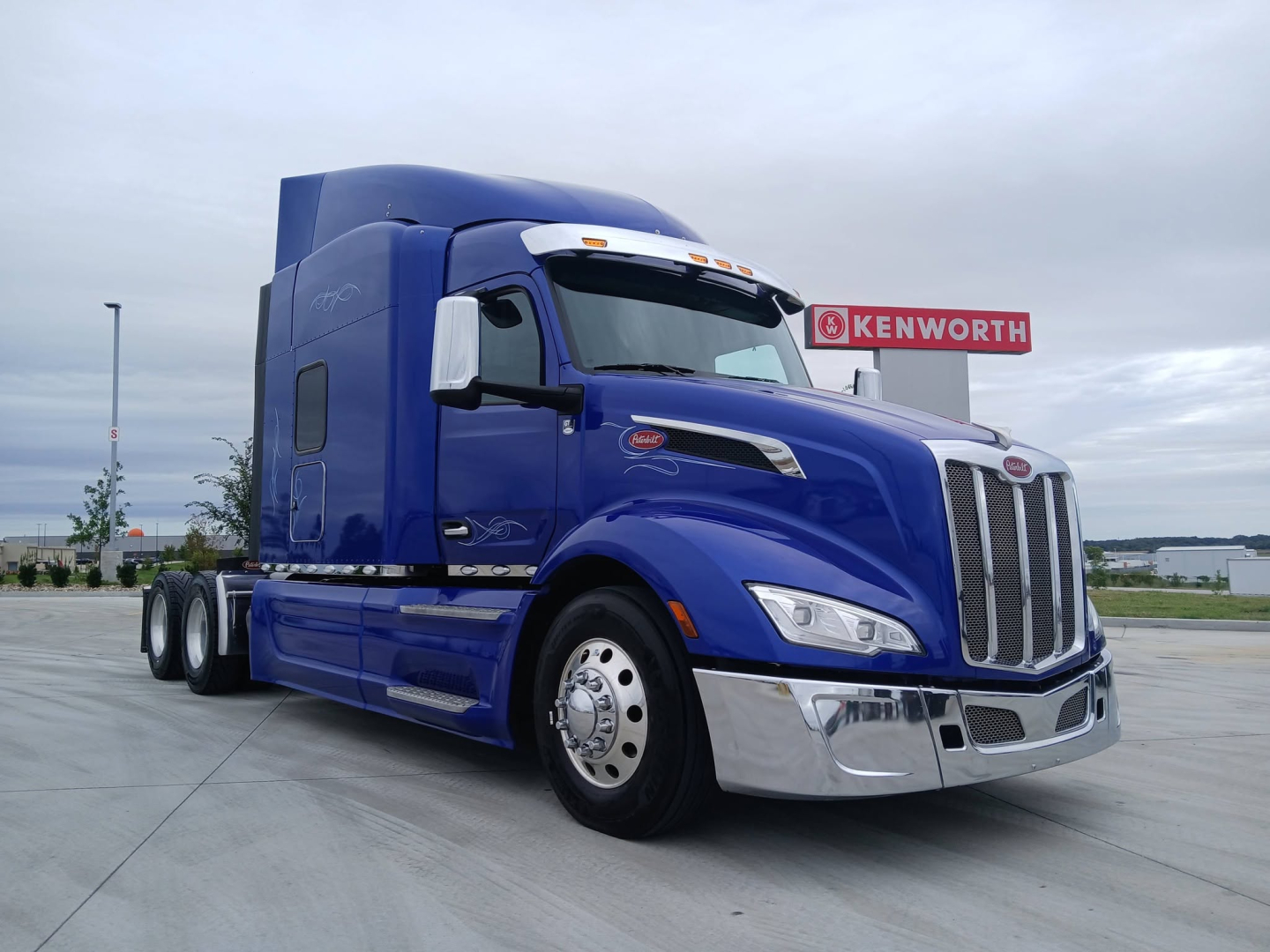 Peterbilt 579 truck in blue, angled front view, chrome grille, Kenworth sign in background. No visible stickers or text on vehicle apart from generic branding.