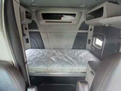 Interior of a Peterbilt 579 sleeper cab showing bed with mattress, overhead storage, and cabin windows.