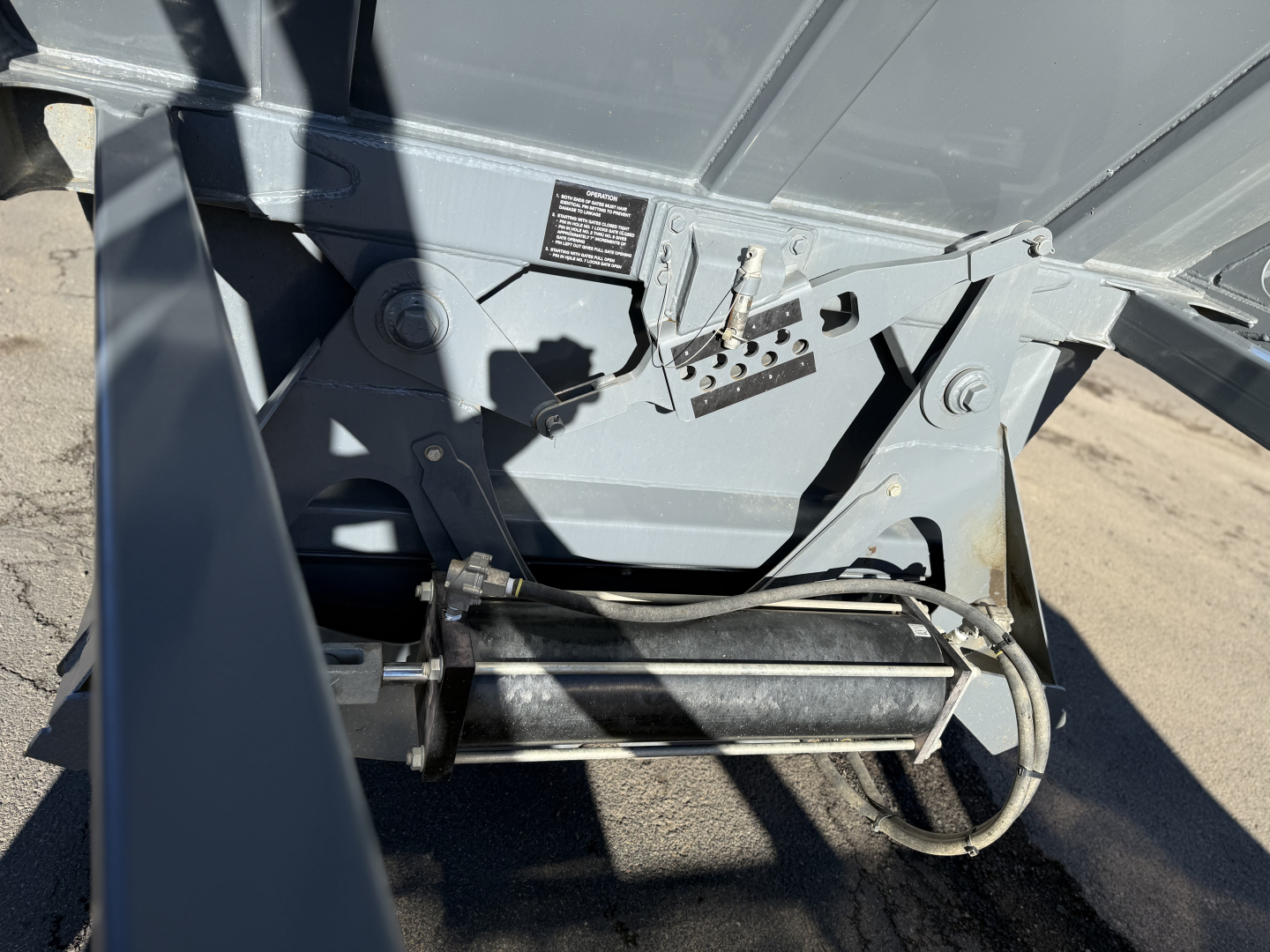 Trailer undercarriage showing a hydraulic pump/roller assembly and control panel; visible label with small text; metal frame and hoses.
