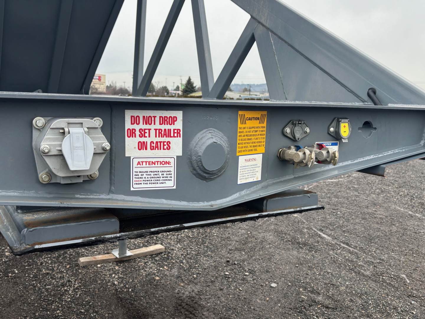 Trailer axle side showing warning labels, hydraulic connections, and support leg; steel frame with Do Not Drop or Set Trailer on Gates sticker and caution placards.