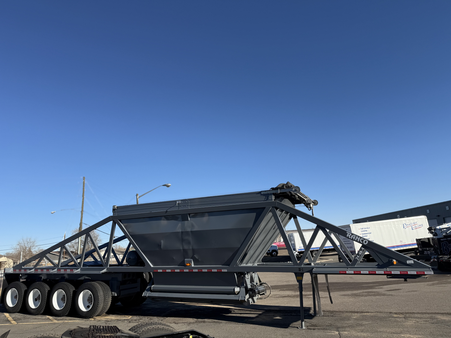 Dump trailer showing LW24464 twin-axle gooseneck frame with large hopper, tri-axle rear, black painted steel construction, clear blue sky.