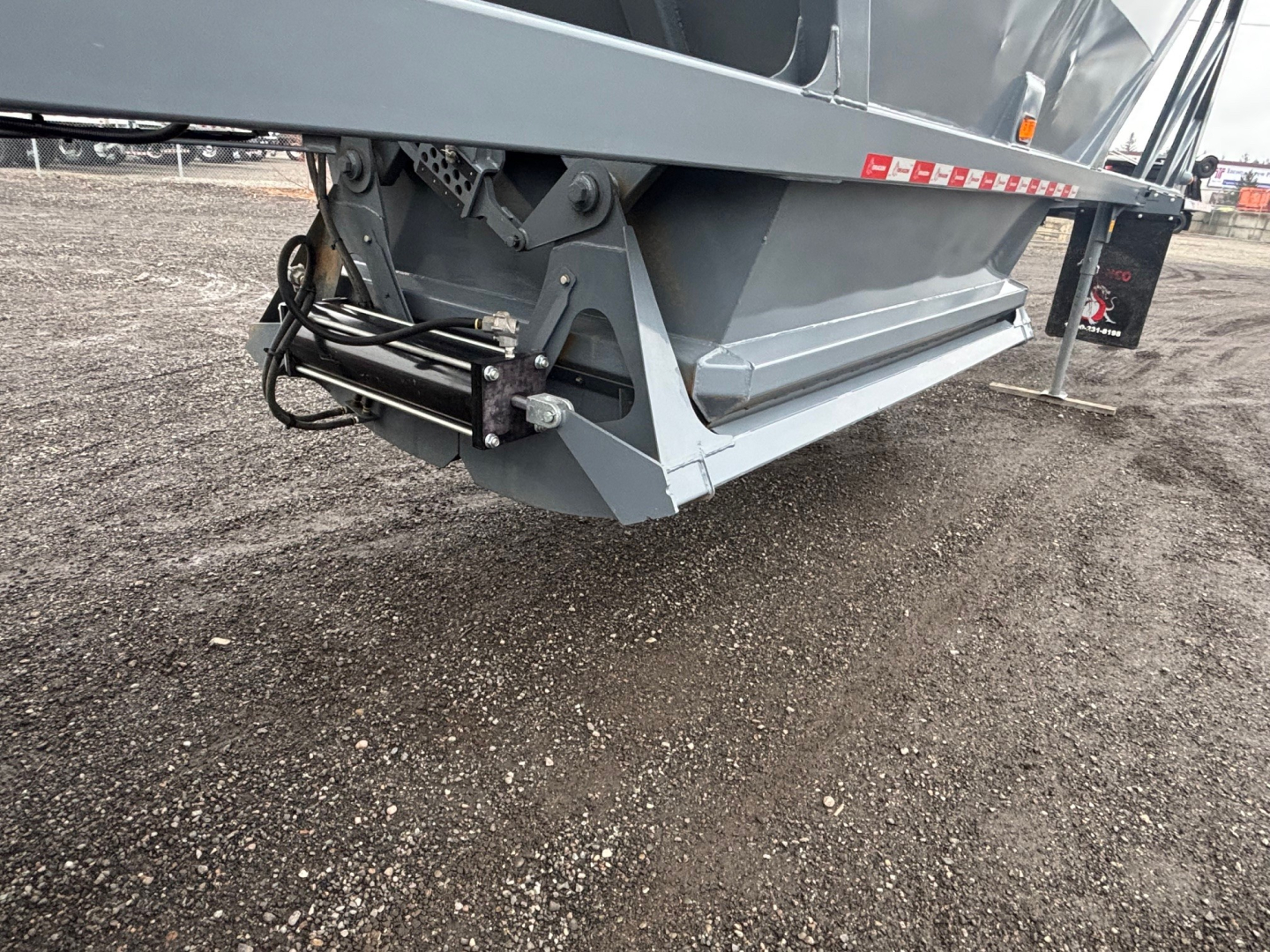 Dump trailer corner showing underside with hydraulic wiring and hitch assembly; gray metal frame and black hoses visible.