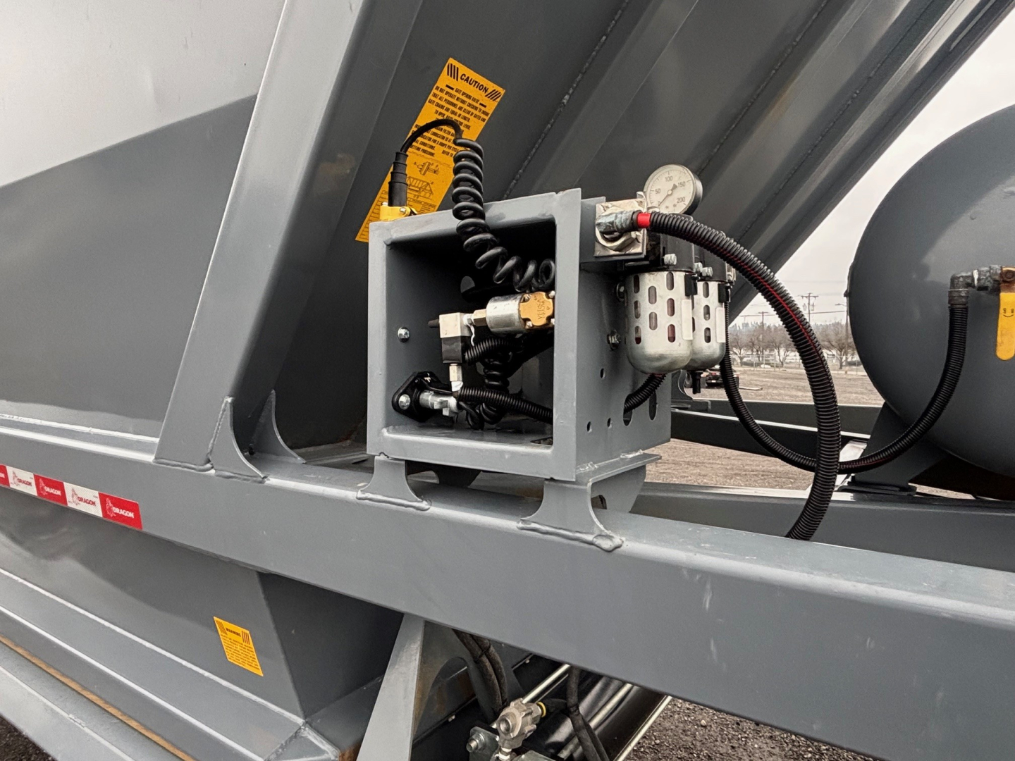 Trailer component platform showing hydraulic pump and control box with hoses and wiring, gray metal frame, warning labels.