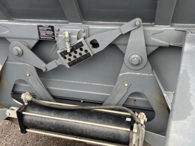 Truck trailer rear mechanism with metal arms and hydraulic cylinder; small black label and a perforated metal block with holes.