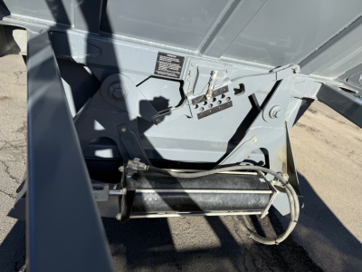 Trailer undercarriage showing a hydraulic pump/roller assembly and control panel; visible label with small text; metal frame and hoses.