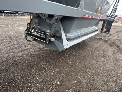 Dump trailer corner showing underside with hydraulic wiring and hitch assembly; gray metal frame and black hoses visible.