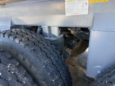 Underframe of a dump trailer showing suspension and dual wheels with tread pattern; label sticker visible on side rail
