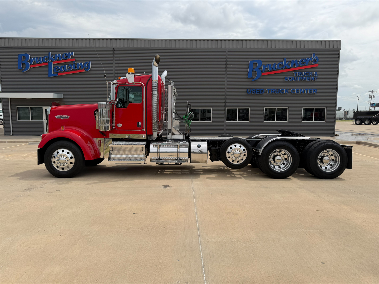 Kenworth W900L conventional daycab truck in red with chrome accents and dual rear axle, parked in front of a building with Buckner Leasing signage.