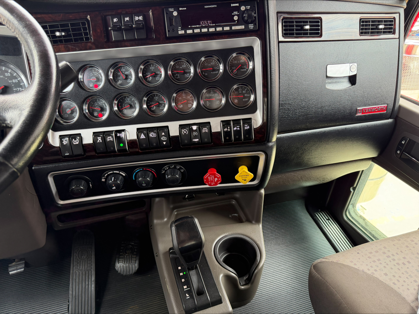 Kenworth W900L dashboard and center console with multiple gauges, control switches, gear selector, and cup holders visible.