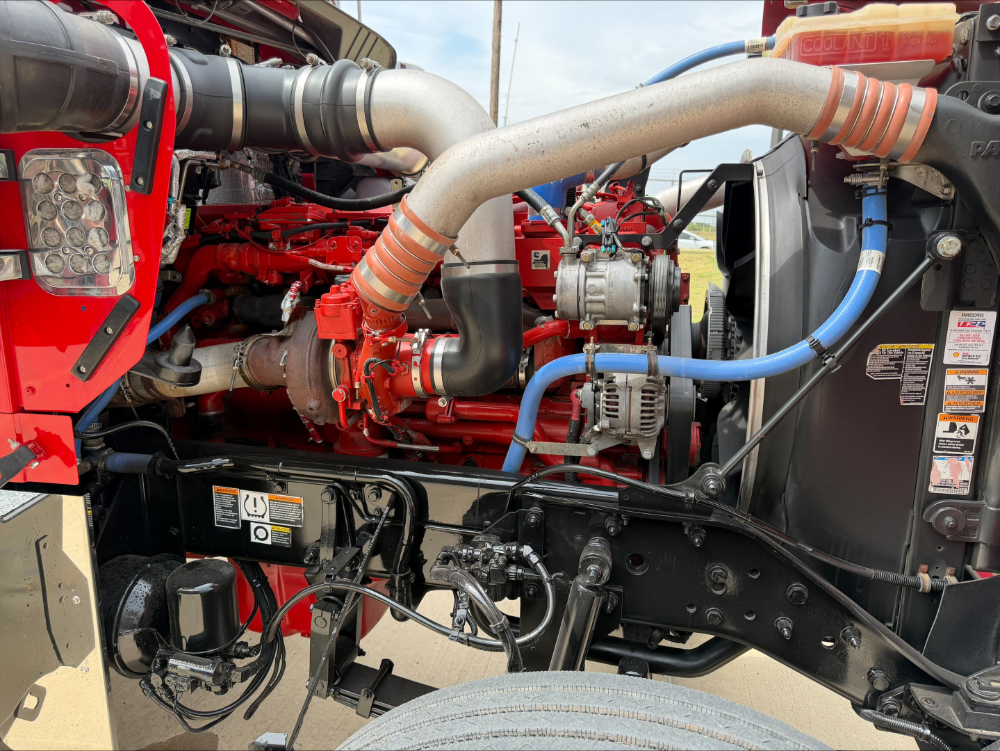 Kenworth W900L engine bay showing red engine block, large intake tubing, aluminum intercooler piping, blue hose, and various warning labels.