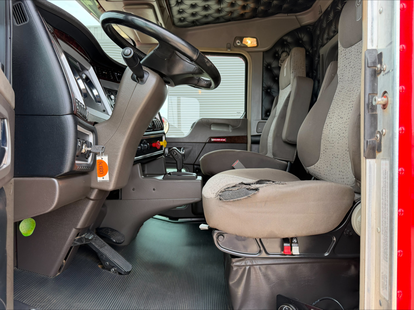 Kenworth W900L truck interior showing driver seat with wear, steering wheel, dashboard, and passenger seat in cab with door open.