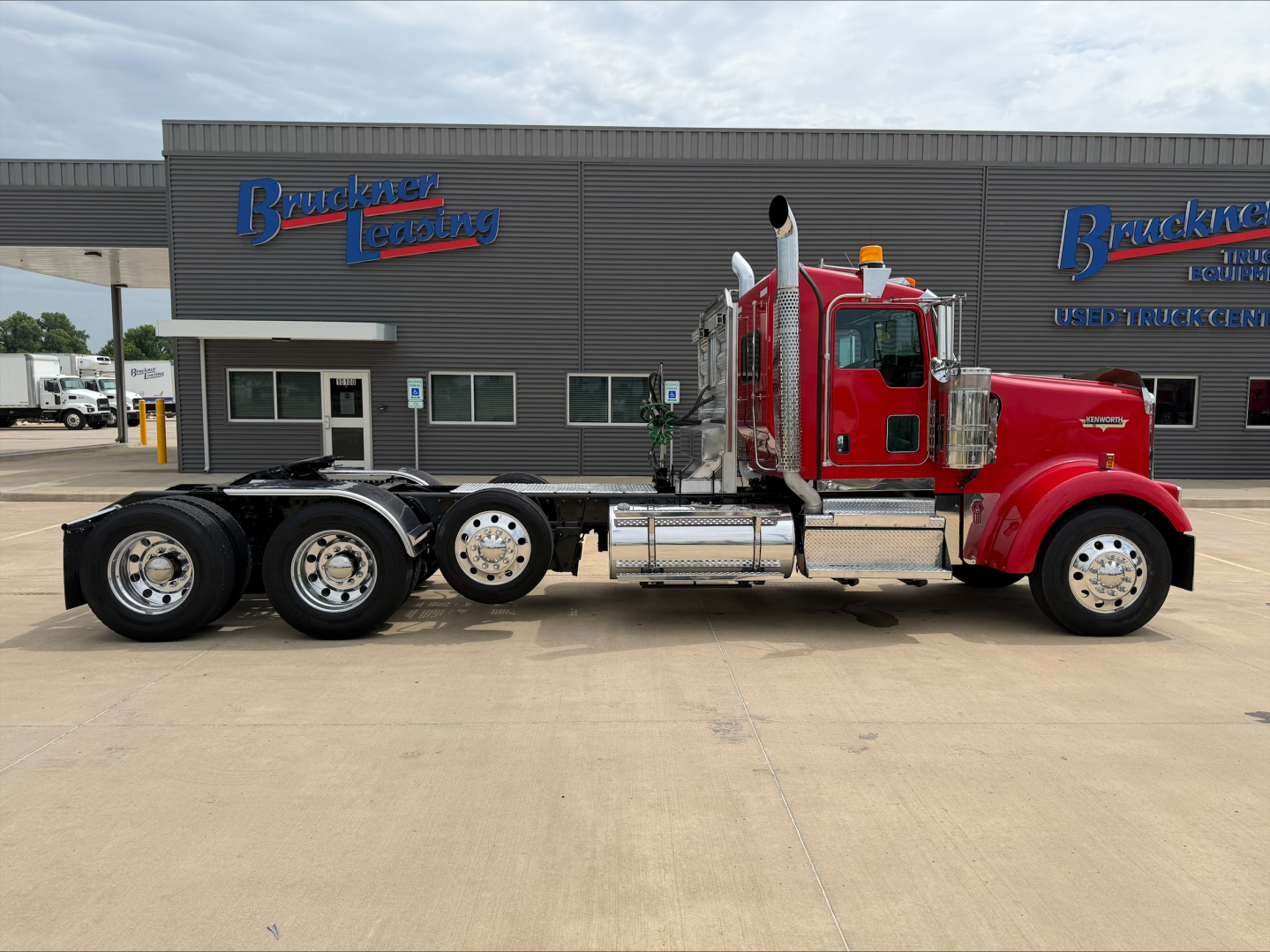 Kenworth W900L tractor side view showing chrome fuel tanks and exhaust stacks, red cab, black dual rear axles, manufacturer sign in background.