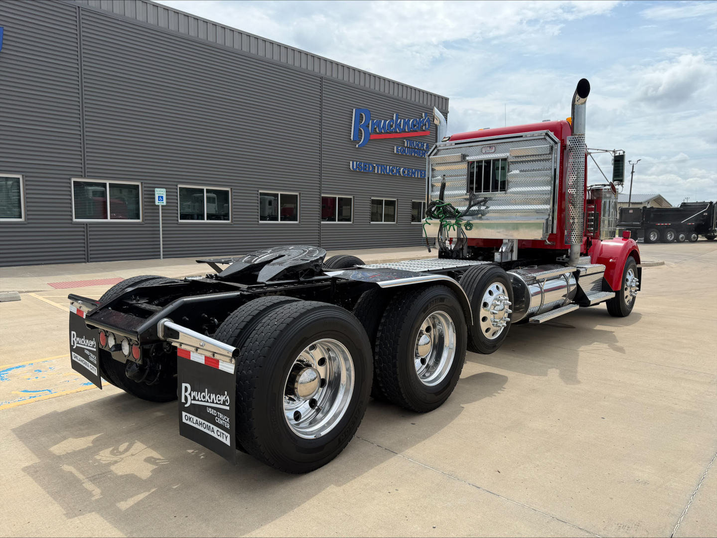 Kenworth W900L tractor in red with chrome fuel tanks and dual rear axles, showcasing truck chassis and cab rump, front of dealership building in background.