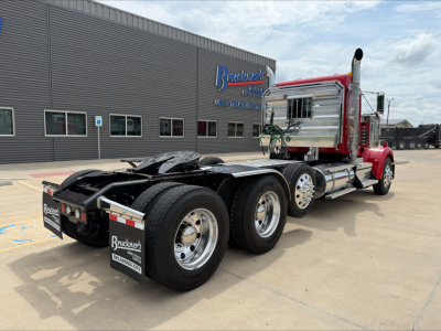 Kenworth W900L tractor in red with chrome fuel tanks and dual rear axles, showcasing truck chassis and cab rump, front of dealership building in background.
