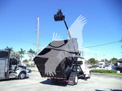 Truck in a dealership lot with a large mechanical lifting hook and grapple attached to a crane over a Peterbilt 337 off a flatbed; visible text includes none clearly.