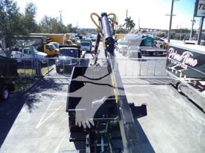 Peterbilt 337 roll-off truck crane arm extending from flatbed with surrounding yard of heavy equipment.