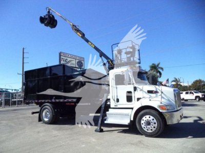 Peterbilt 337 rolloff truck with crane in raised position, side view, white cab, black hopper, visible hitch and stabilizer, dealership lot in background.