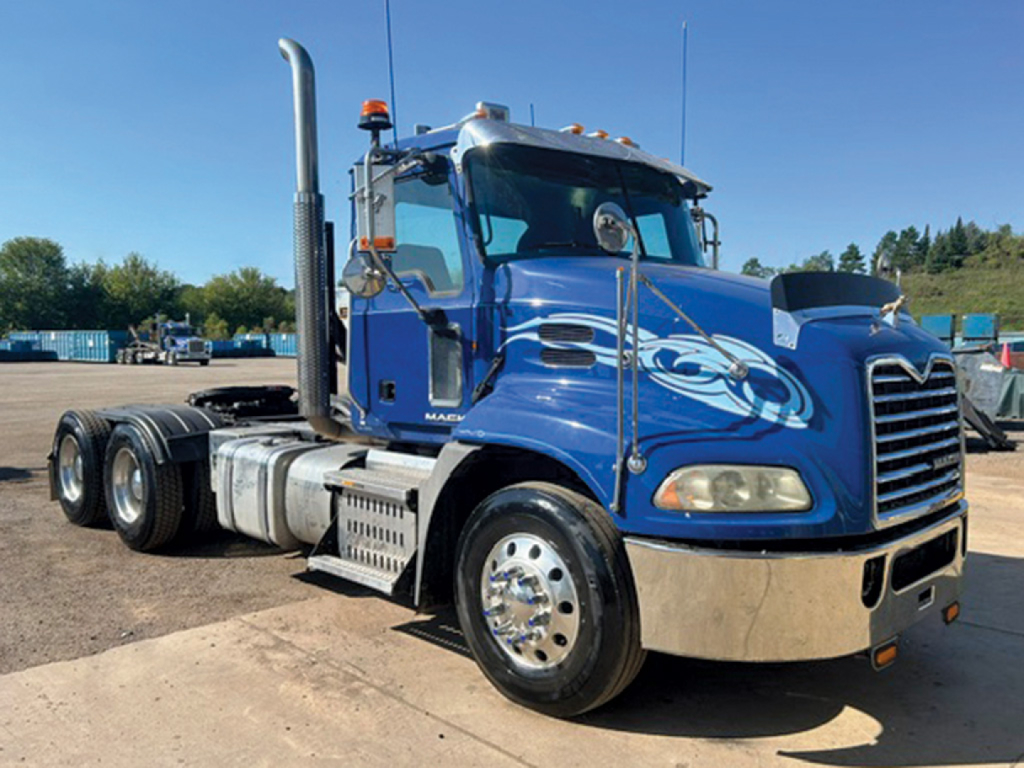 Truck cab and front half of a blue 2014 Mack CXU613 conventional daycab showing chrome bumper, exhaust stack, and hood with decorative graphic.