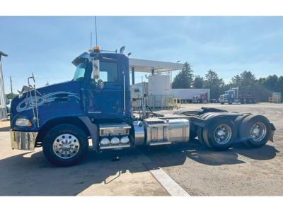 Blue 2014 Mack CXU613 daycab tractor in service showing chrome fuel tanks and rear triple axle configuration.